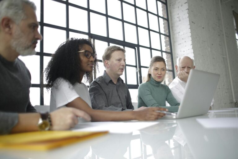 From below of focused multiracial colleagues of different ages in casual wear surfing netbook while making project together in light workplace